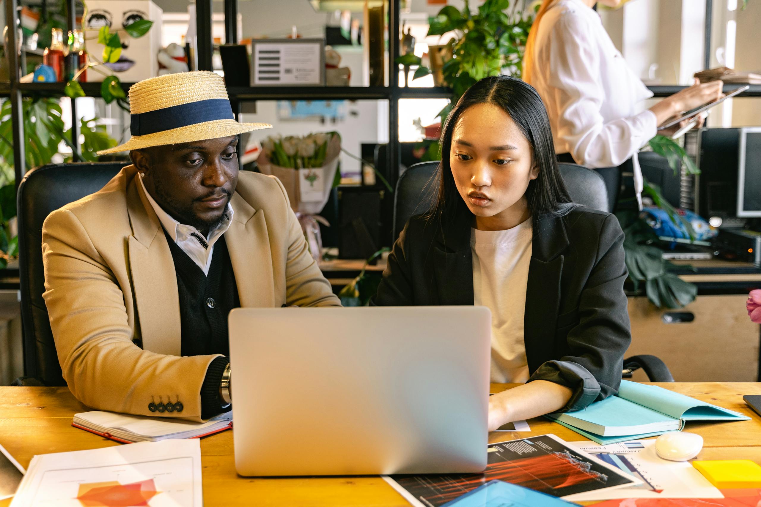 Asian woman and black man engaged in collaboration at a modern office setting, working on a laptop.