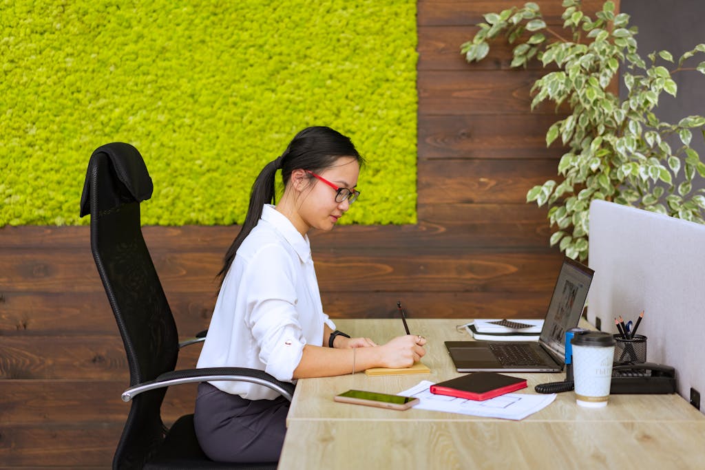 Asian woman in office setting, focused on writing notes at her desk.