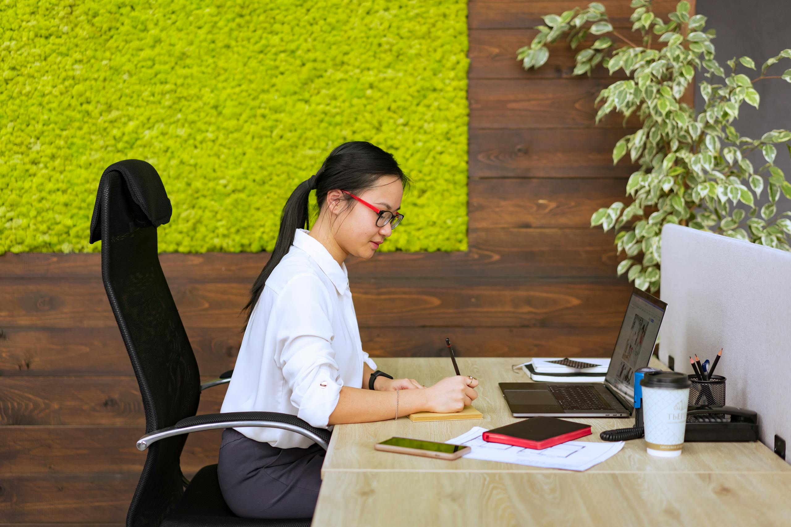 Asian woman in office setting, focused on writing notes at her desk.