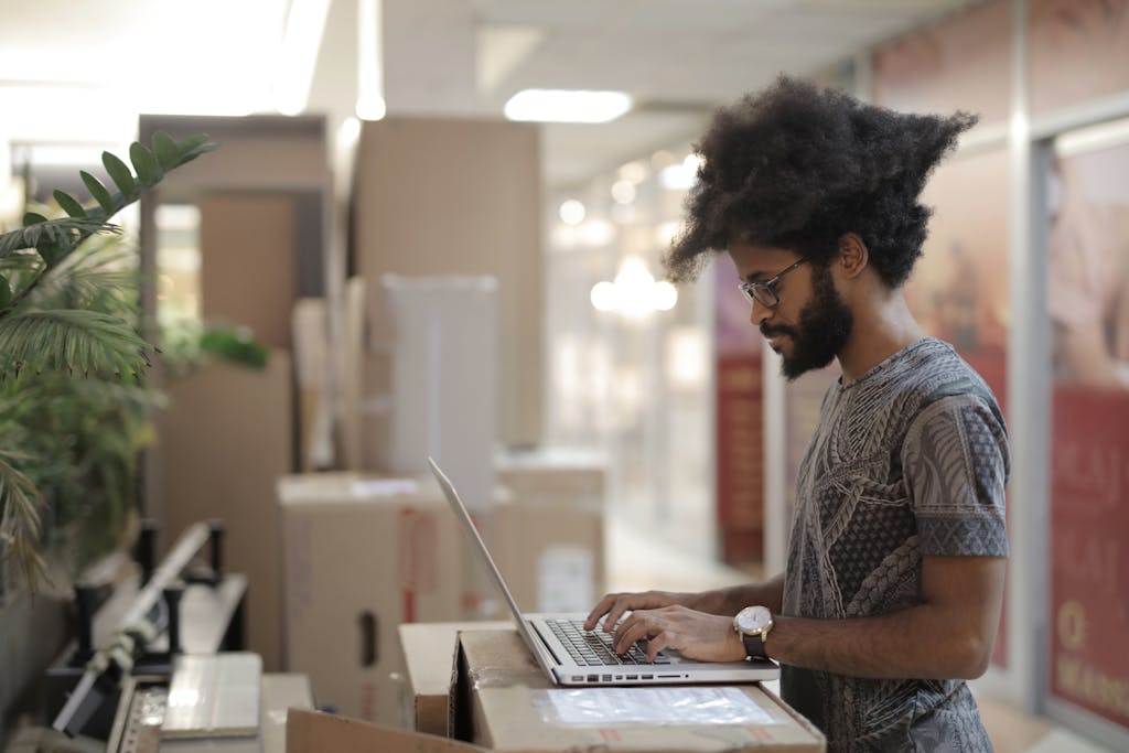 Side view of ethnic male with Afro hairstyle and in casual clothes with eyeglasses using laptop and thinking while working on project in modern workspace